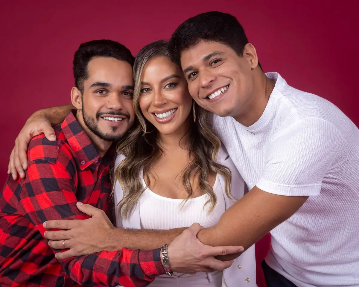 Três pessoas abraçadas diante de um fundo vermelho, com roupas casuais; destaque para camisa xadrez vermelha e preta, peça branca e camiseta clara, além de acessórios metálicos no pulso.