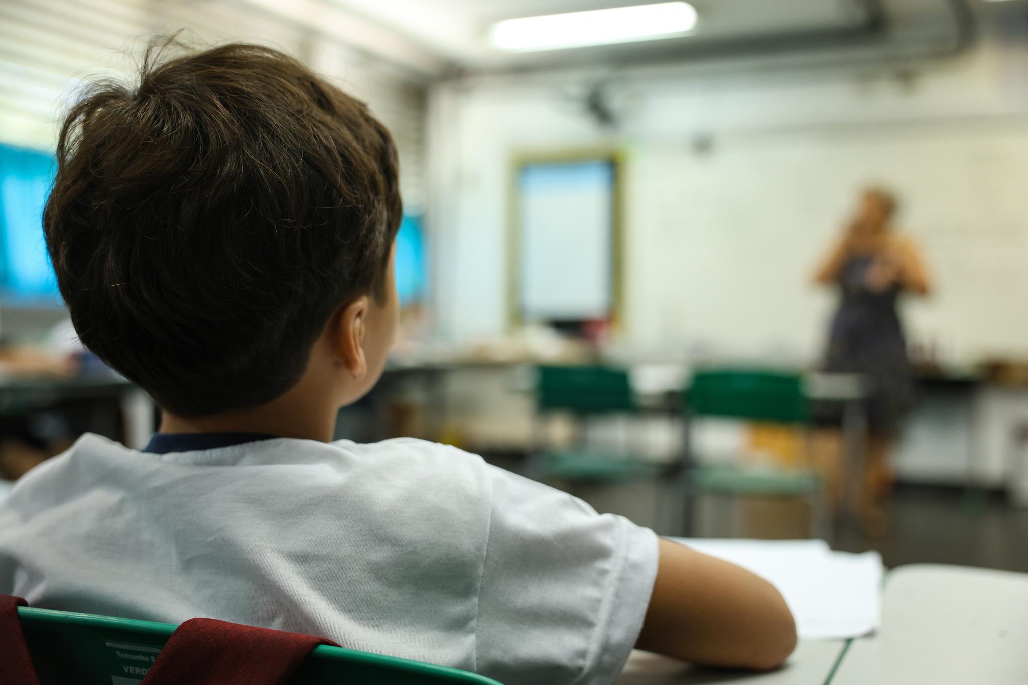 Foto ilustrativa de criança em sala de aula, de costas, olhando para a professora que está dando aula.