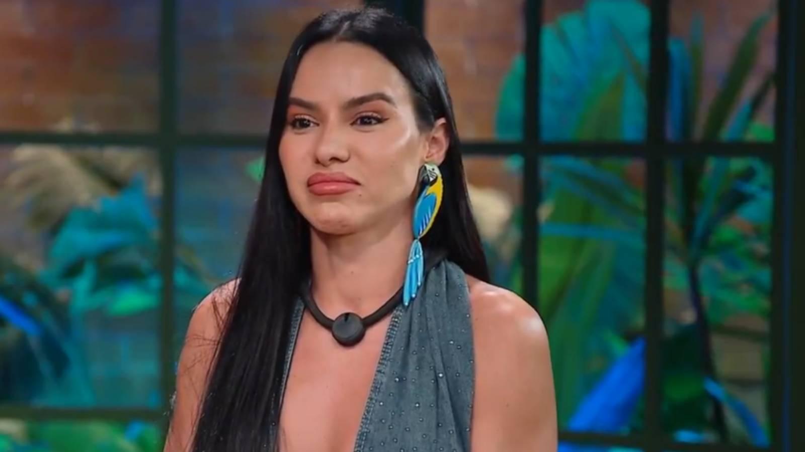 Imagem mostra a participante do reality show A Fazenda 17, Tàmires Assîs, em close-up, com top azul-acinzentado, colar preto e feição que denota tristeza ou desapontamento, após ser eliminada na 11ª roça do programa.