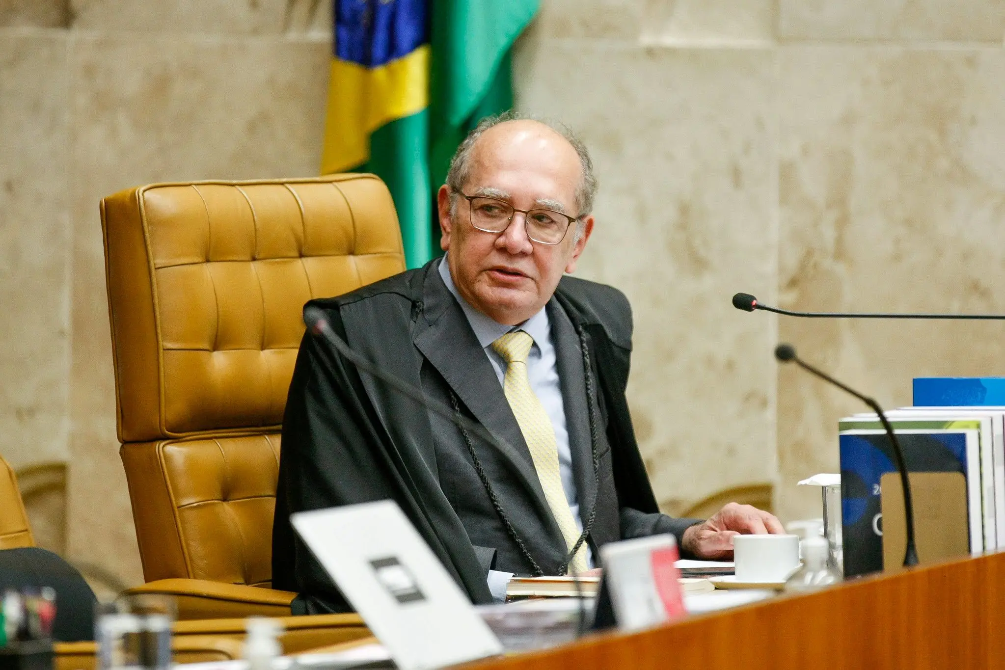 Ministro do Supremo Tribunal Federal, Gilmar Mendes, em sessão plenária, usando toga e óculos, com a bandeira do Brasil ao fundo.