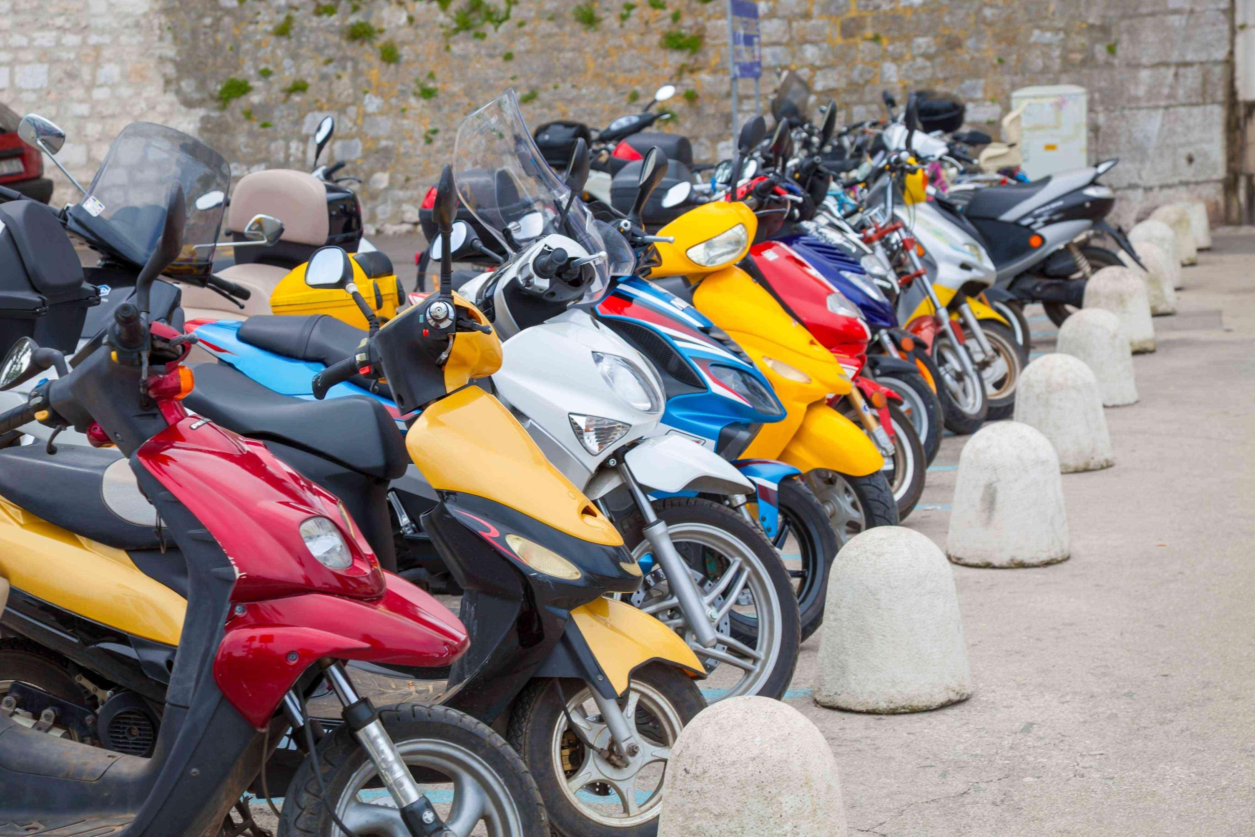 Imagem mostra sequência de scooters, tipo ciclomotor, e motocicletas em várias cores estacionadas ao longo de uma rua com barreiras de pedra.