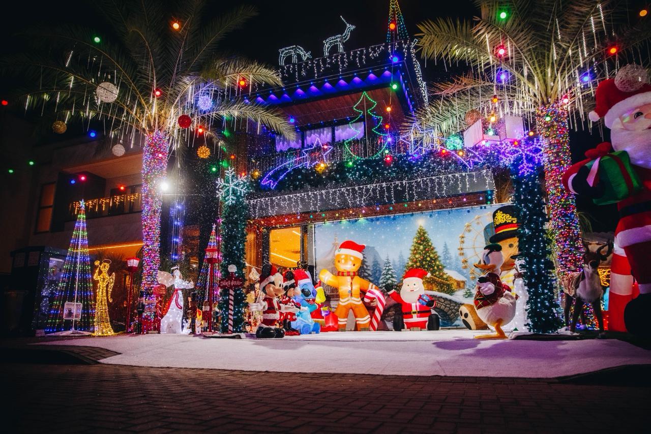Esta é uma fotografia noturna e muito colorida de uma casa intensamente decorada para o Natal. O tema principal é de iluminação exuberante e grandes infláveis temáticos.
No centro da cena, a fachada da casa é coberta por milhares de luzes piscantes de cores variadas (vermelho, verde, azul, amarelo), com destaque para luzes azuis e brancas penduradas que simulam gelo ou chuva de estrelas. Há várias silhuetas de cervos no telhado, também iluminadas.
Em frente à casa, há uma grande coleção de bonecos infláveis de Natal e personagens de desenhos animados que preenchem o primeiro plano. Eles estão dispostos sobre o que parece ser um tapete branco que simula neve.