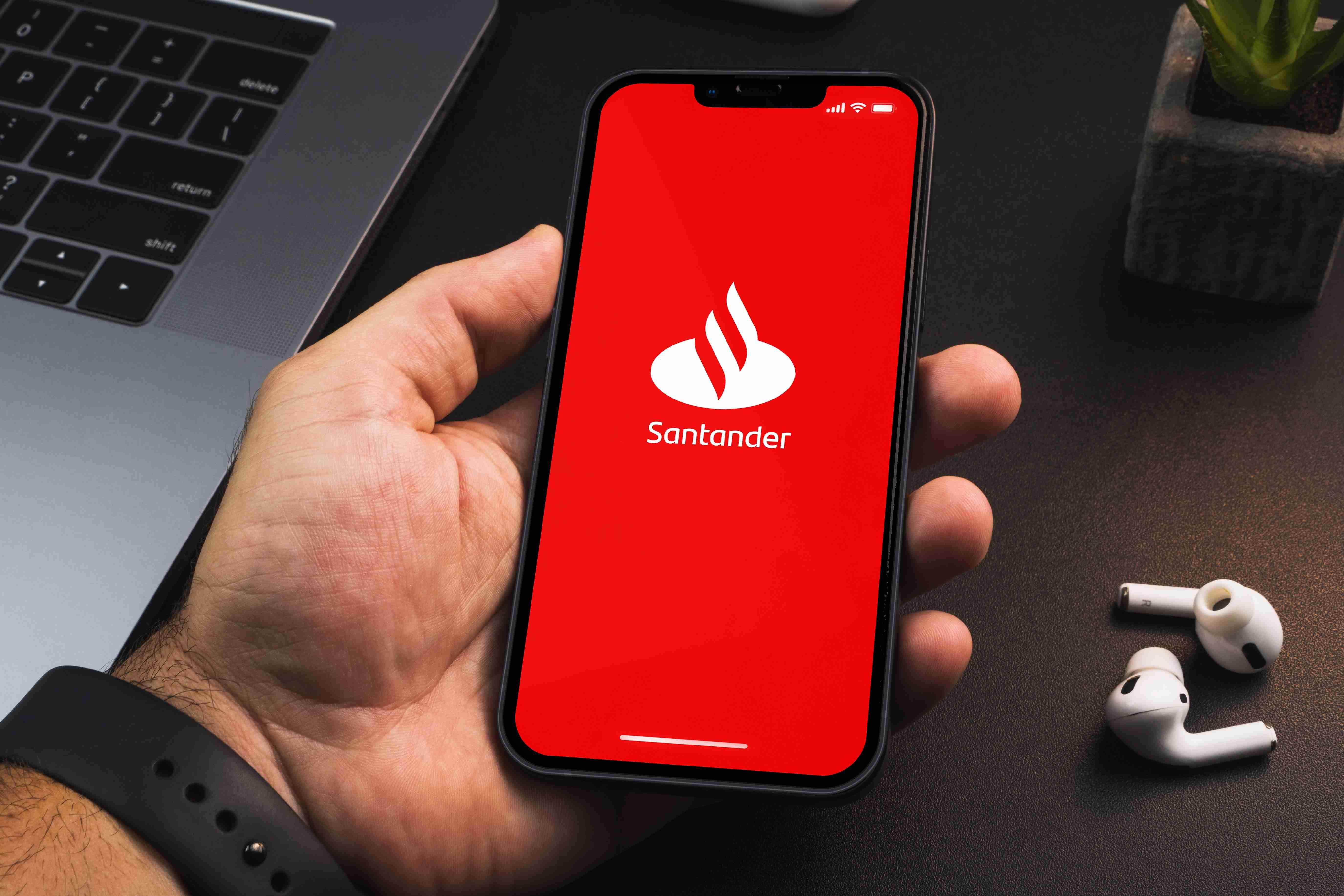 Imagem mostra mão segurando um smartphone com o aplicativo do Banco Santander aberto em sua tela vermelha.