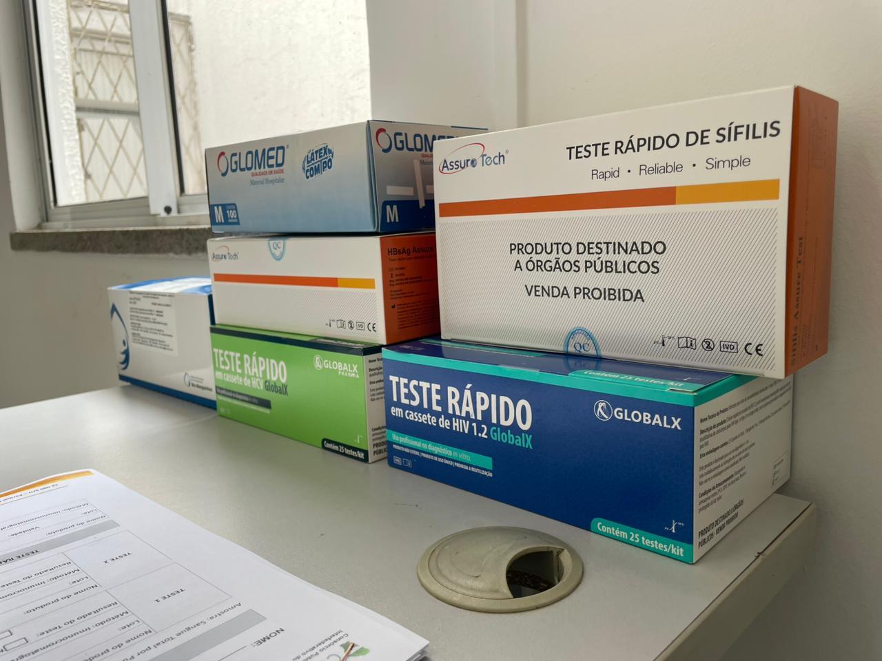 Imagem mostra caixas de testes rápidos para HIV empilhadas sobre uma mesa de um equipamento de saúde.
