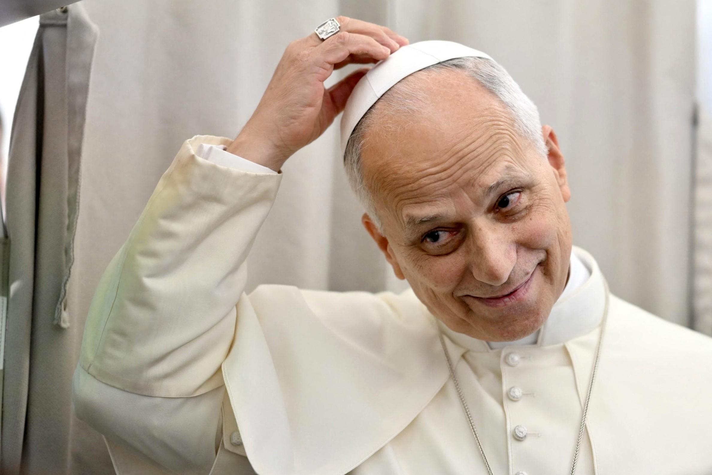 Imagem mostra papa Leão XIV sorrindo e ajustando seu solidéu branco com a mão direita, onde um anel proeminente é visível.