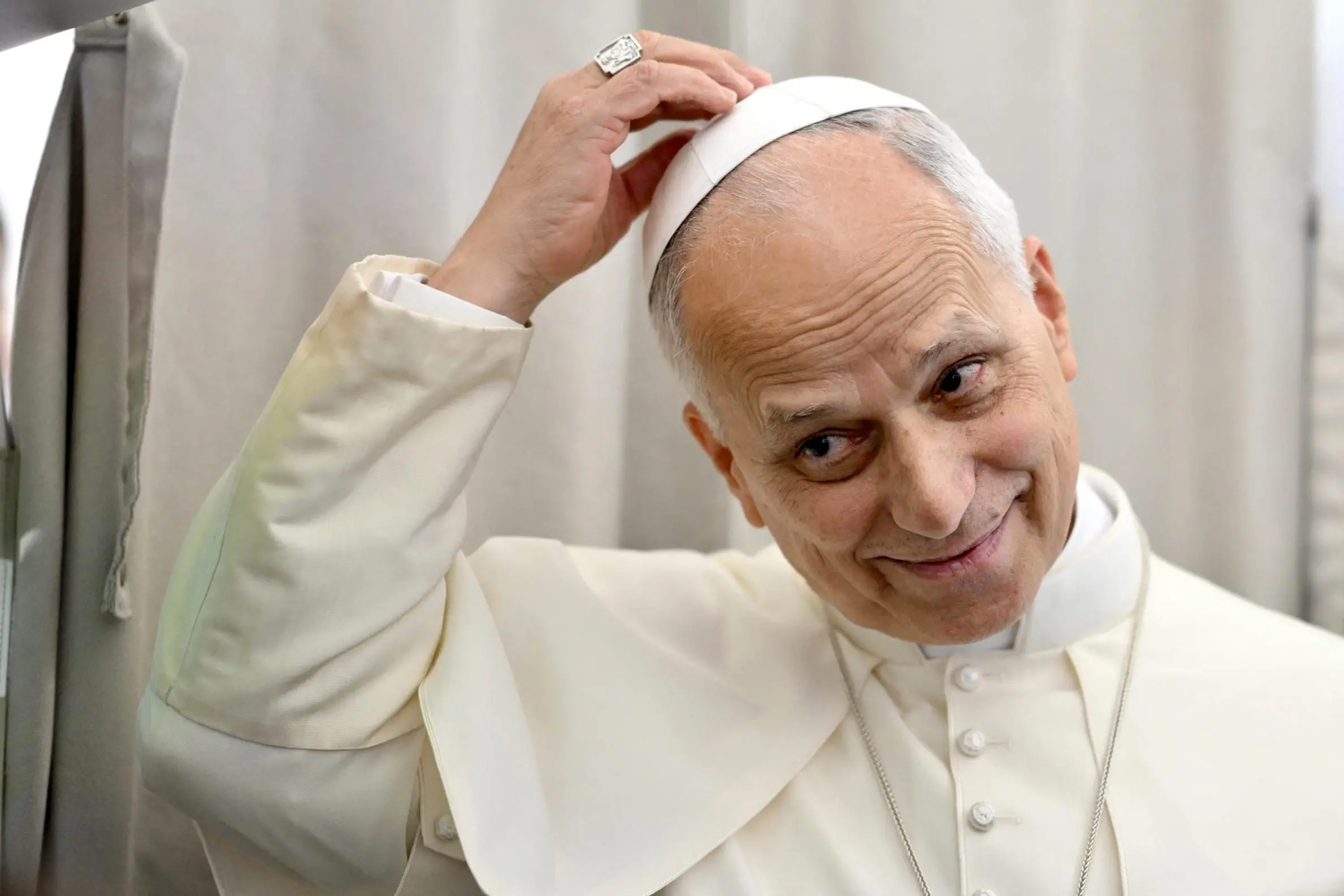 Imagem mostra papa Leão XIV sorrindo e ajustando seu solidéu branco com a mão direita, onde um anel proeminente é visível.