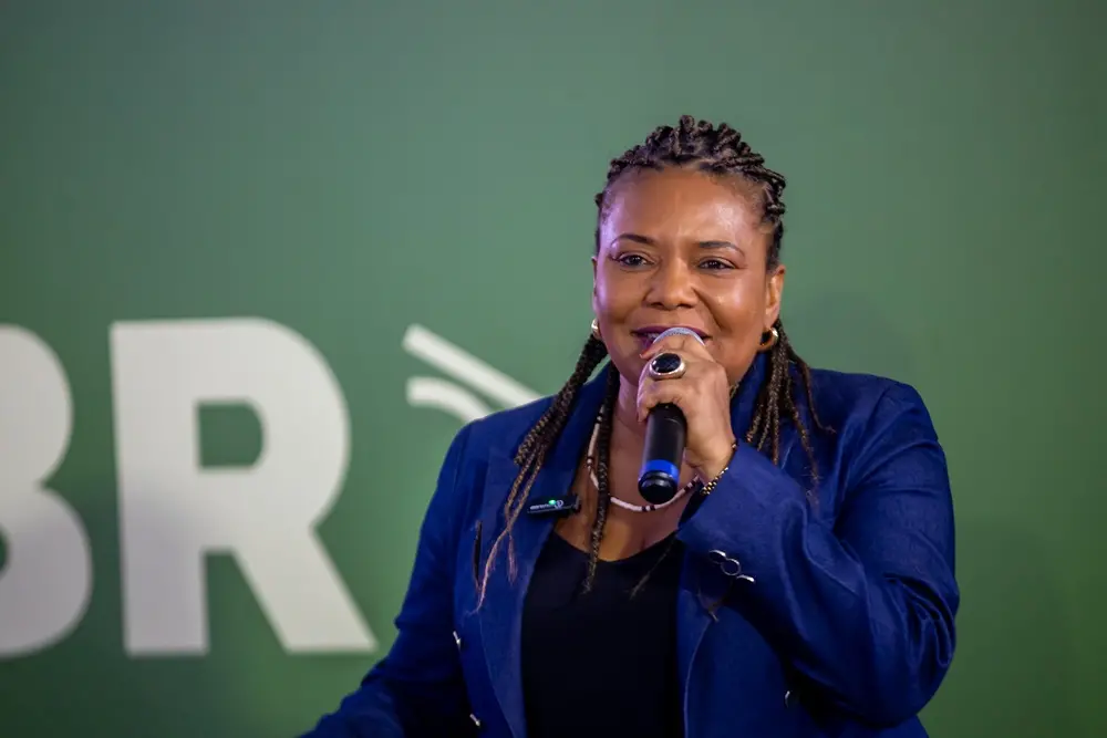 Na imagem, uma mulher de pele escura, com cabelo trançado, sorri enquanto segura um microfone com a mão direita perto da boca, aparentemente falando em um evento. Ela veste um blazer azul-marinho de tecido que parece ser jeans ou brim e uma blusa preta por baixo. A mulher usa um anel grande e escuro no dedo anelar direito e brincos de argola dourados. O fundo é de uma cor verde escura e suave, com as letras brancas 