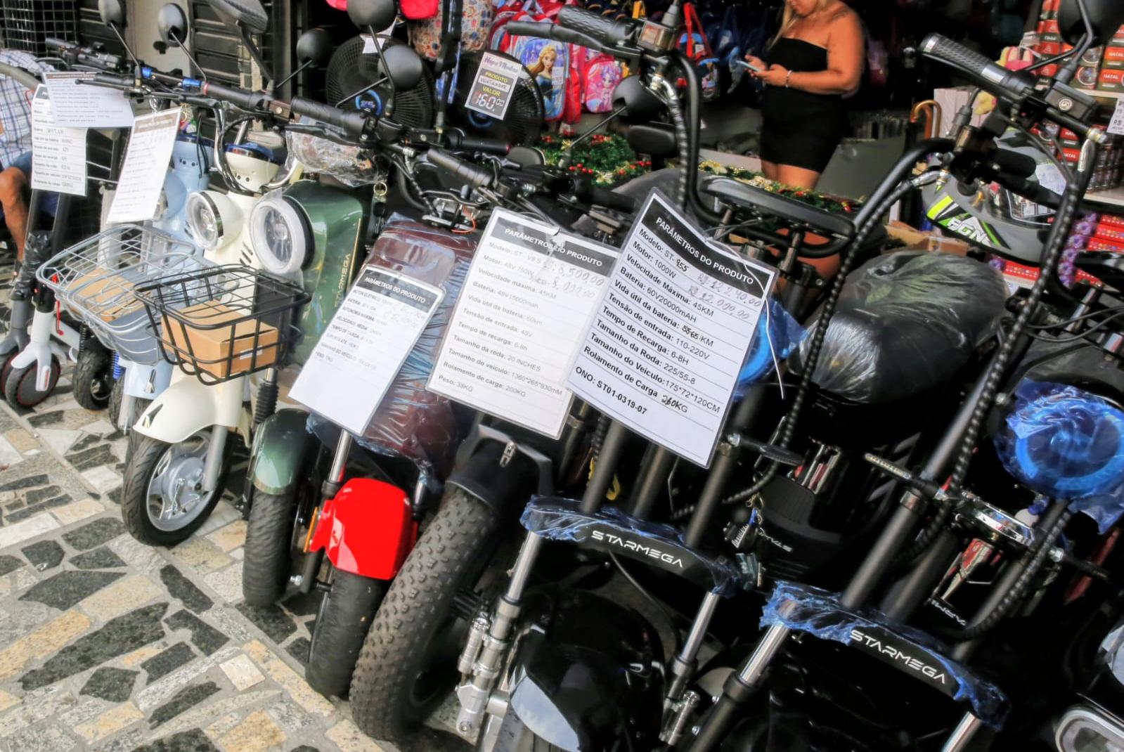 Uma fileira de scooters e ciclomotores elétricos estacionados em uma calçada, com etiquetas de preço visíveis. Uma mulher em pé no fundo do lado direito.