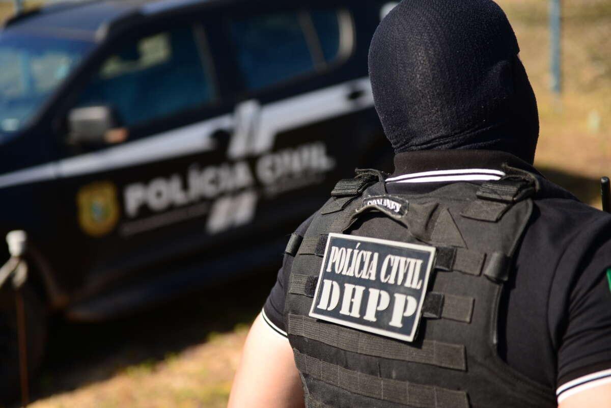 A imagem mostra um policial civil do DHPP, fardado, com balaclava e colete balístico, e uma viatura da Polícia Civil ao fundo.