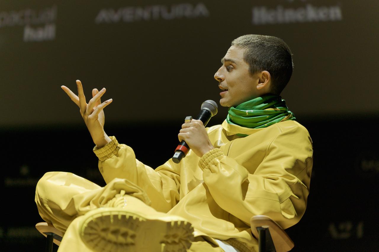 Timothée Chalamet em passagem por evento de Marty Supreme no Brasil com camisa amarela e bandeira do Brasil.