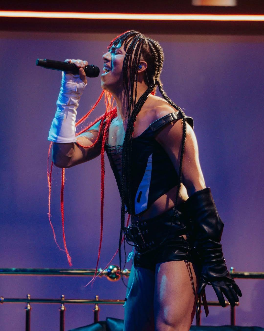 Artista em show ao vivo, com expressão intensa, cantando em microfone sob luzes roxas no palco.