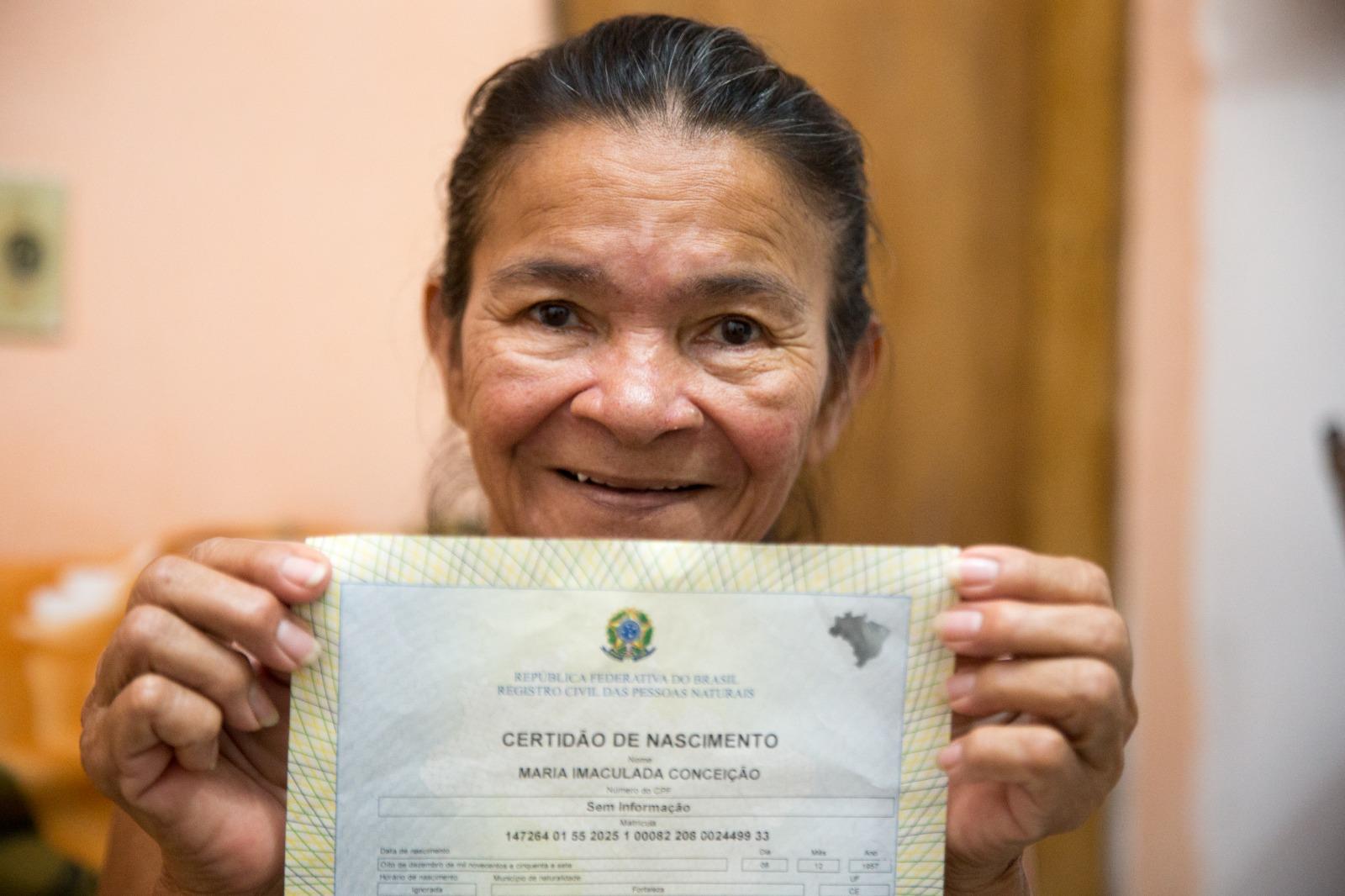 Na imagem, retrato em close-up de uma mulher brasileira mais velha, de pele morena, sorrindo e olhando diretamente para a câmera. Ela segura, com as duas mãos, um documento oficial do governo brasileiro, uma Certidão de Nascimento (Certidão de Nascimento), diretamente em frente ao peito. A parte visível do documento está centrada e mostra o título 