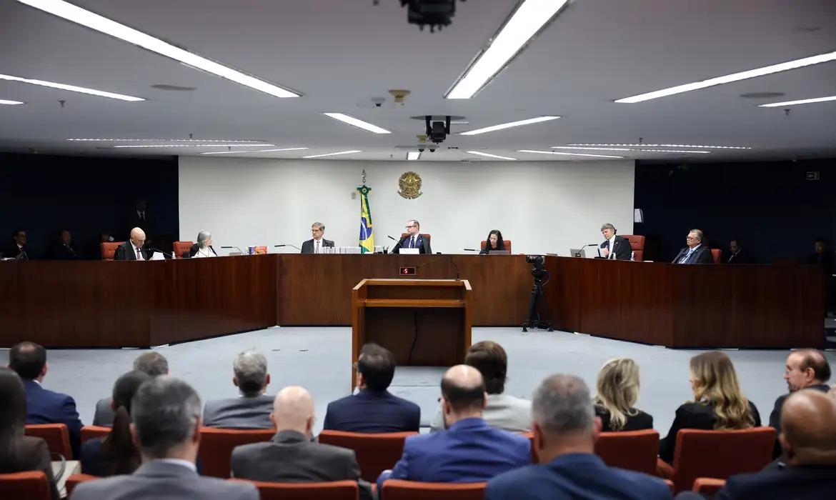 Sala de julgamento com ministros e ministras sentados em uma bancada semicircular de madeira. No centro, há um púlpito vazio voltado para o plenário. Ao fundo, vê-se a bandeira do Brasil e o brasão da República na parede. Diversas pessoas assistem à sessão sentadas nas primeiras fileiras, enquanto câmeras registram o momento.