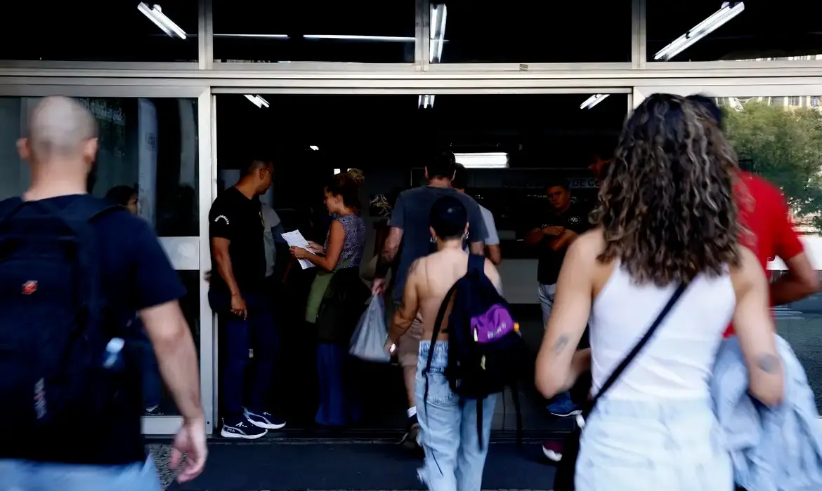 Pessoas entram em um prédio público por uma porta de vidro aberta; algumas carregam mochilas, sacolas e documentos. O local parece movimentado, com um grupo reunido próximo à entrada e iluminação fluorescente no interior.