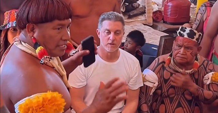 Luciano Huck de camiseta branca está sentado entre dois líderes indígenas com pinturas corporais e ornamentos tradicionais; um deles fala segurando um celular, enquanto outras pessoas da comunidade aparecem ao fundo em um ambiente de aldeia.