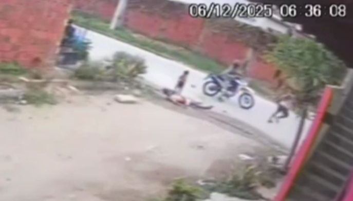 Imagem de câmera de segurança mostra uma rua onde uma pessoa está caída no chão, enquanto duas outras estão próximas; uma delas está ao lado de uma motocicleta parada e outra corre pela calçada. A cena ocorre em área residencial com muros de tijolos e vegetação.
