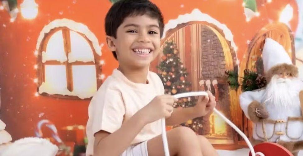 Caso Benício: Criança feliz posando em estúdio temático de Natal, com fundo de casa decorada e figura de Papai Noel.