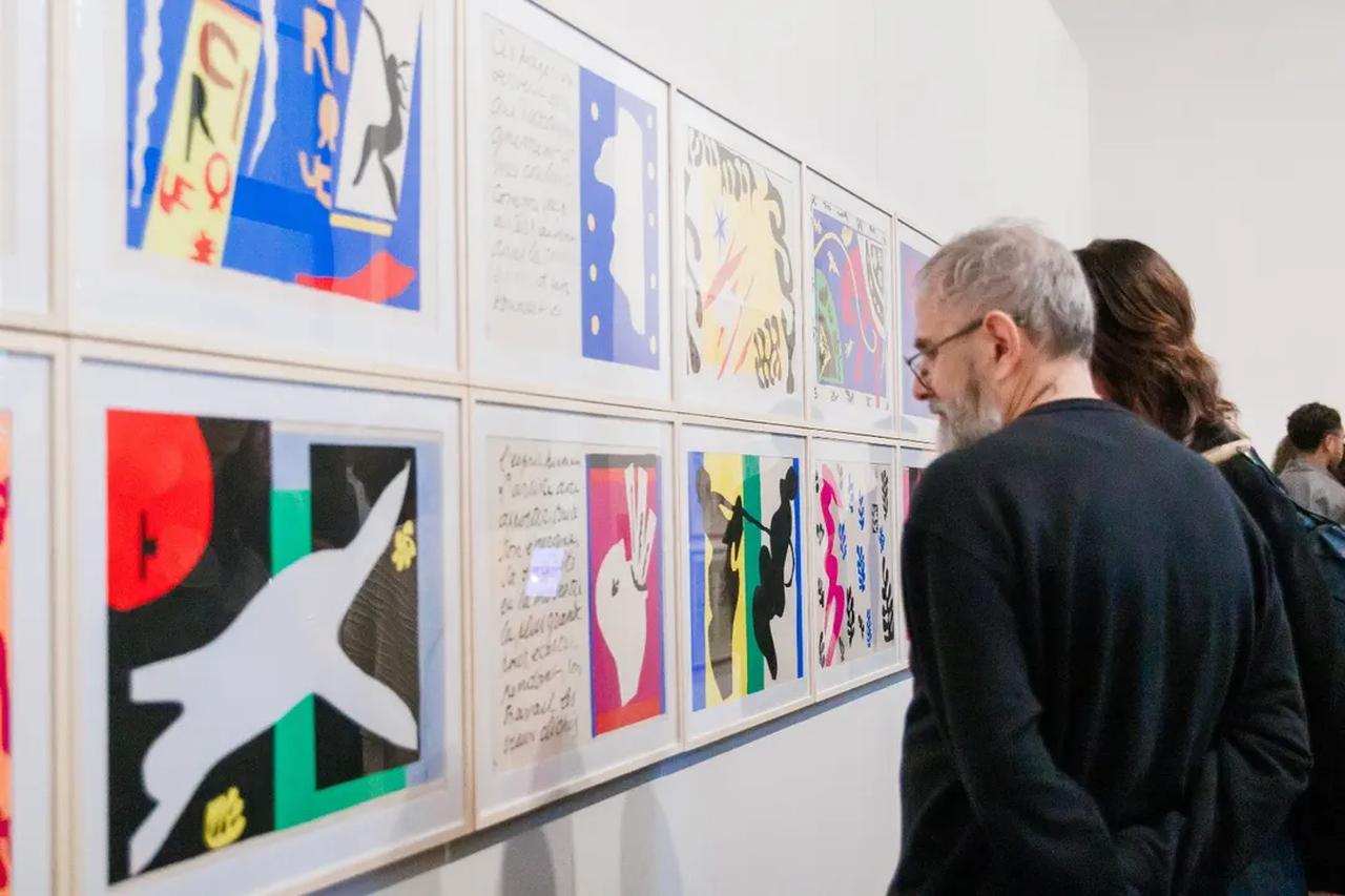 Pessoas olham gravuras penduradas em parede de exposição.