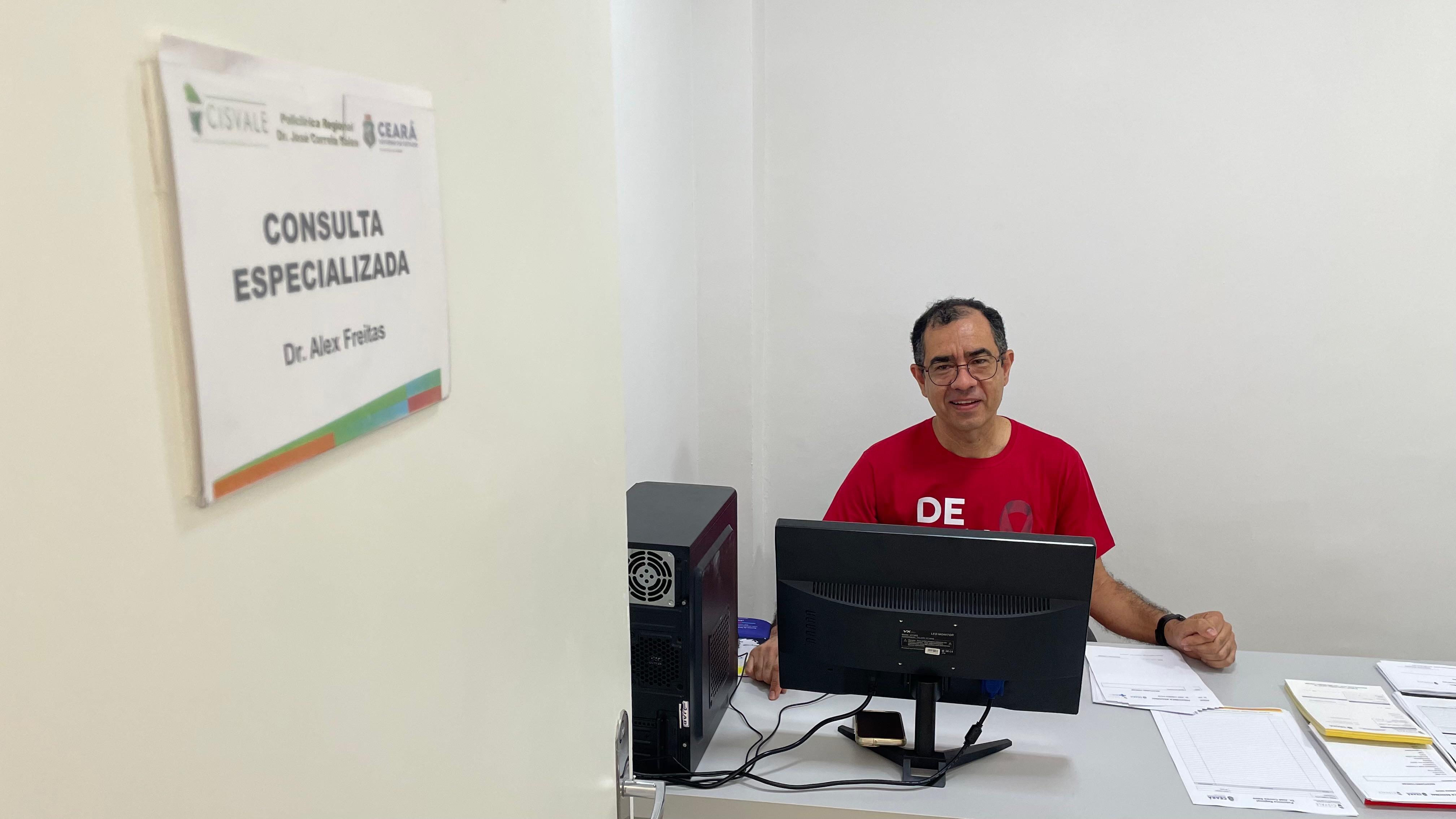 O Dr. Alex Freitas, médico infectologista, aparece sentado em sua mesa, usando uma camisa vermelha, em frente a um monitor de computador. Uma placa na parede identifica o local como Consulta Especializada - Dr. Alex Freitas, na Policlínica Regional de Caucaia.