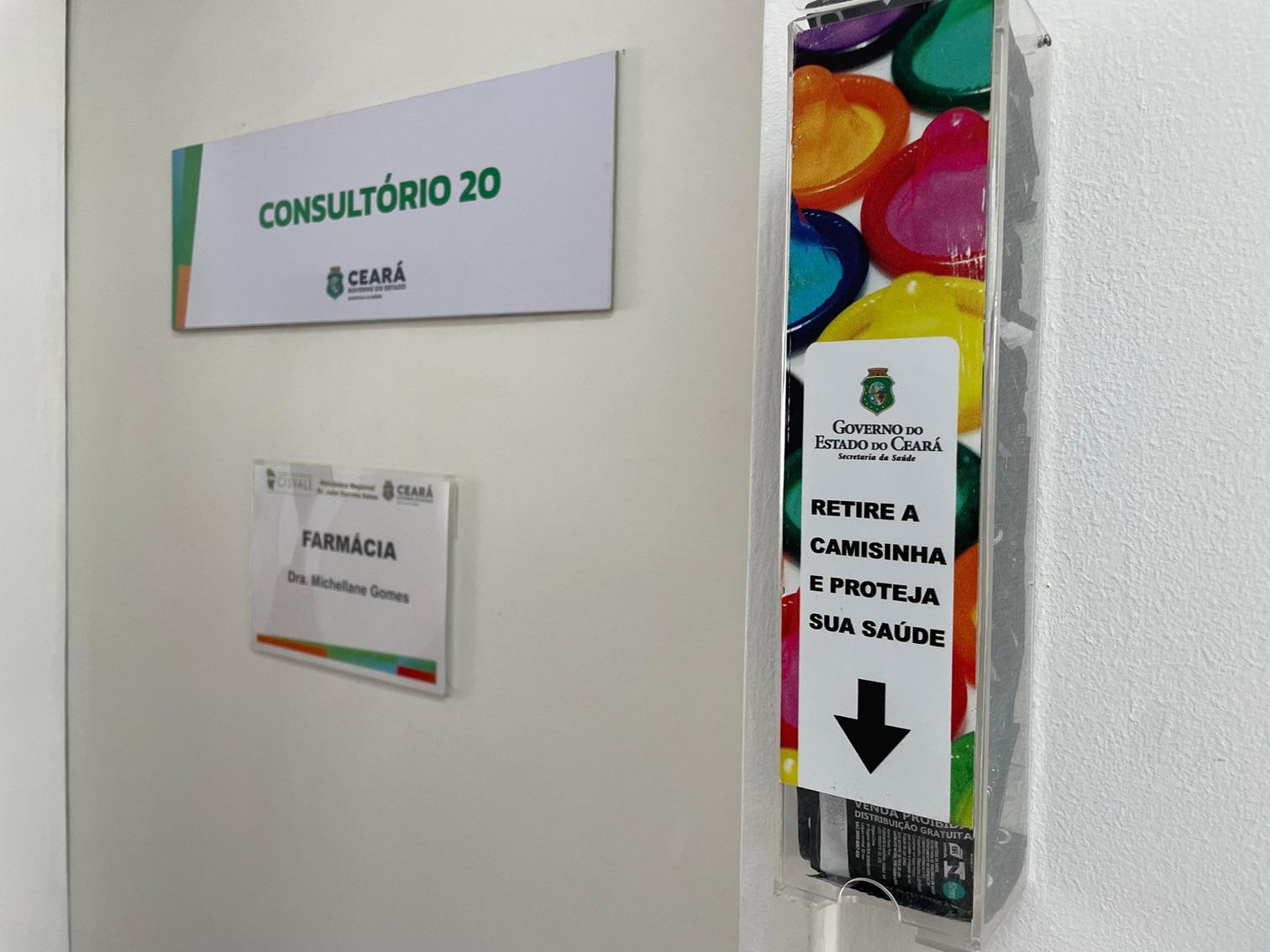 Imagem de um dispensador de camisinhas coloridas fixado em uma parede de um posto de saúde. Ele exibe uma mensagem do Governo do Ceará incentivando a proteção.