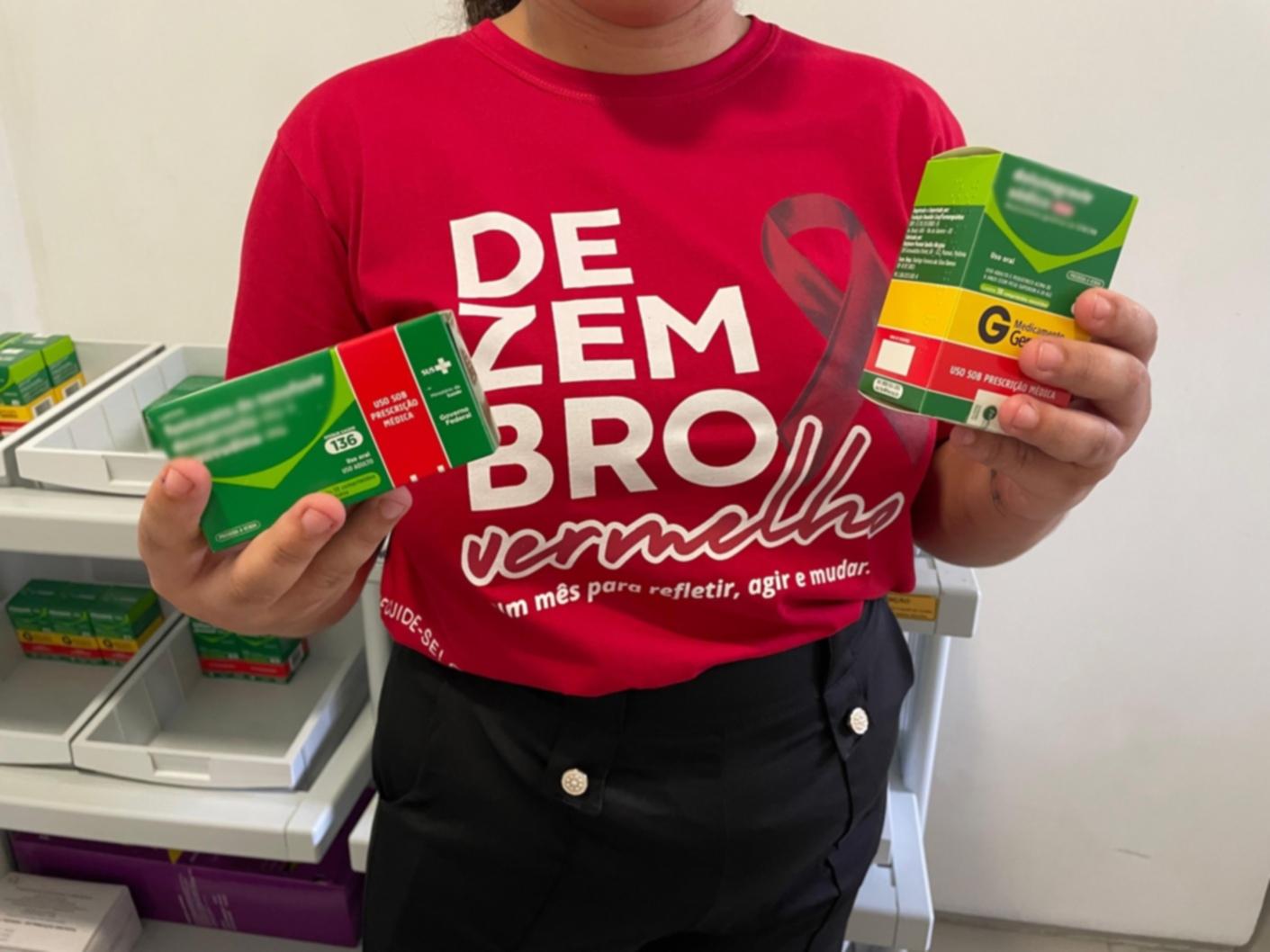 Uma enfermeira, vestindo uma camisa vermelha com a inscrição Dezembro Vermelho, segura duas caixas de medicamentos para HIV. As caixas são verdes e vermelhas, e há prateleiras com mais medicamentos ao fundo.