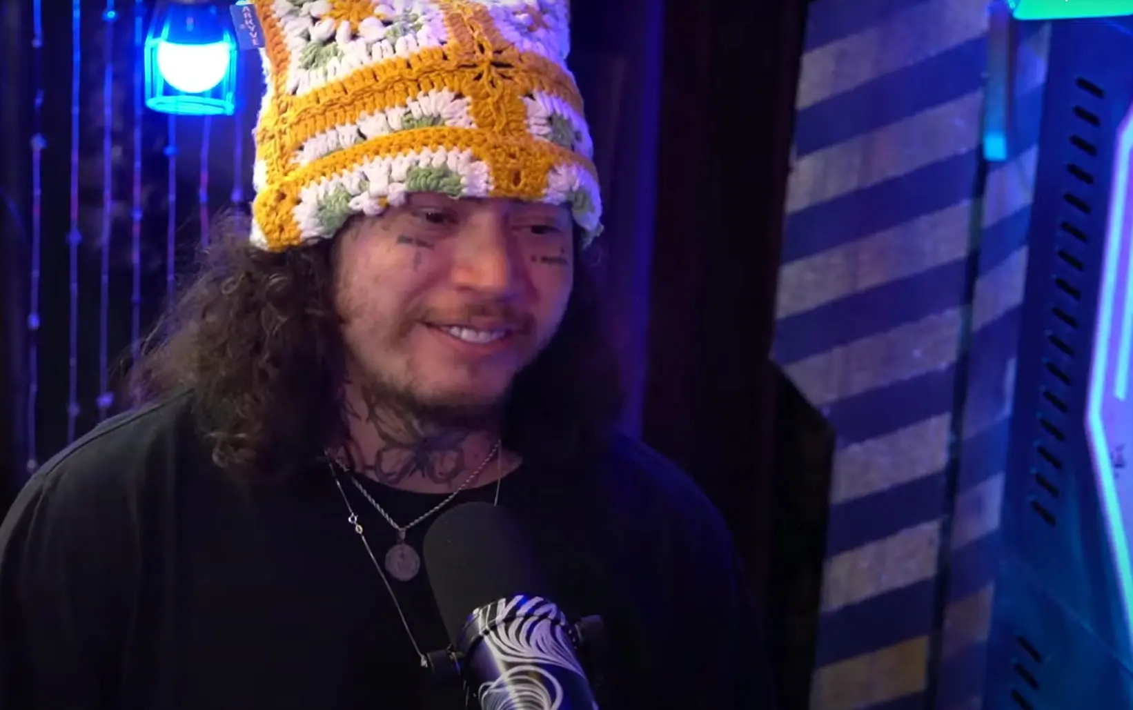 Whindersson Nunes de gorro de crochê colorido e tatuagens faciais, sorrindo em um estúdio de gravação.