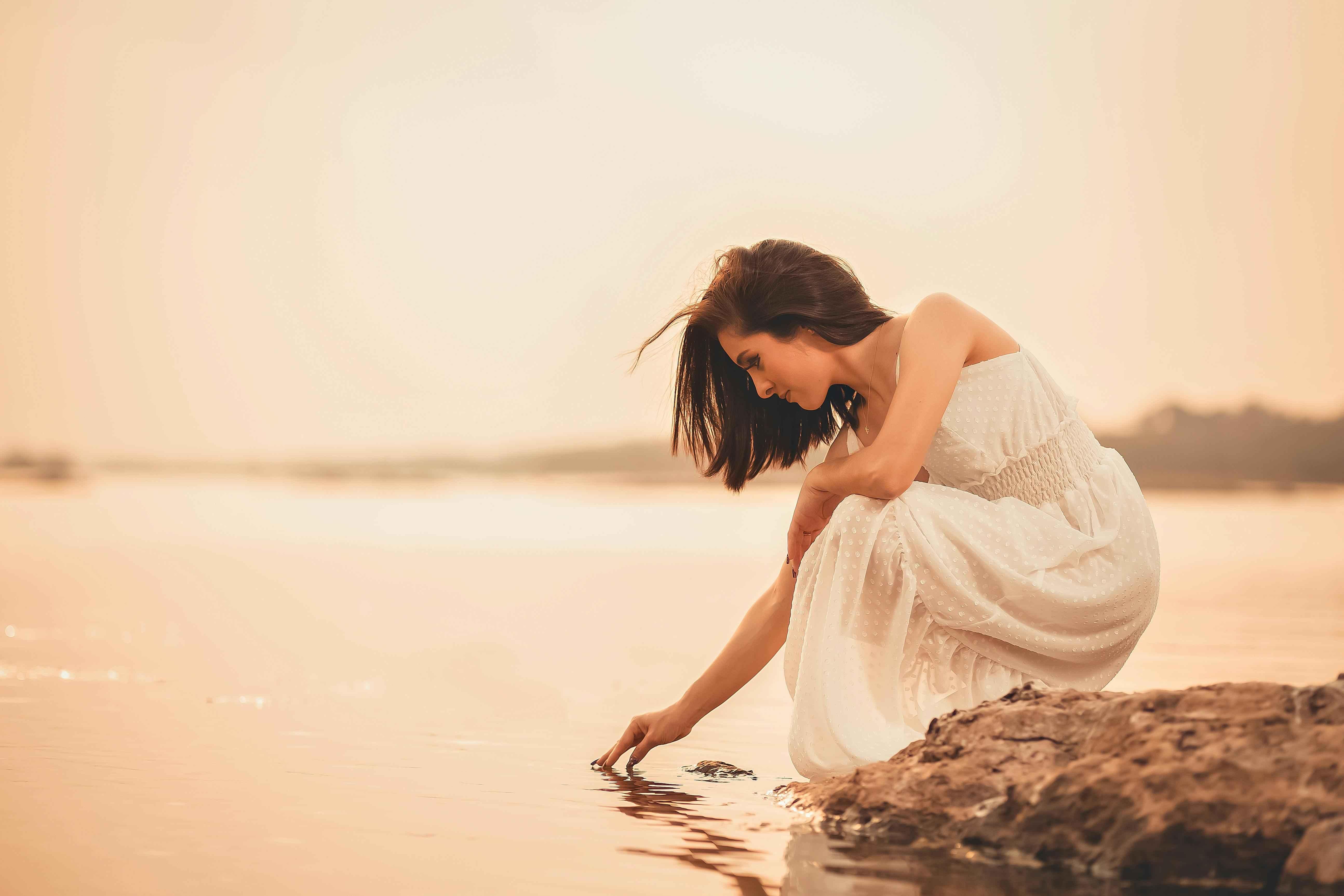 Mulher de vestido branco agachada à beira da água, tocando a superfície com a ponta dos dedos sob um céu de pôr do sol.