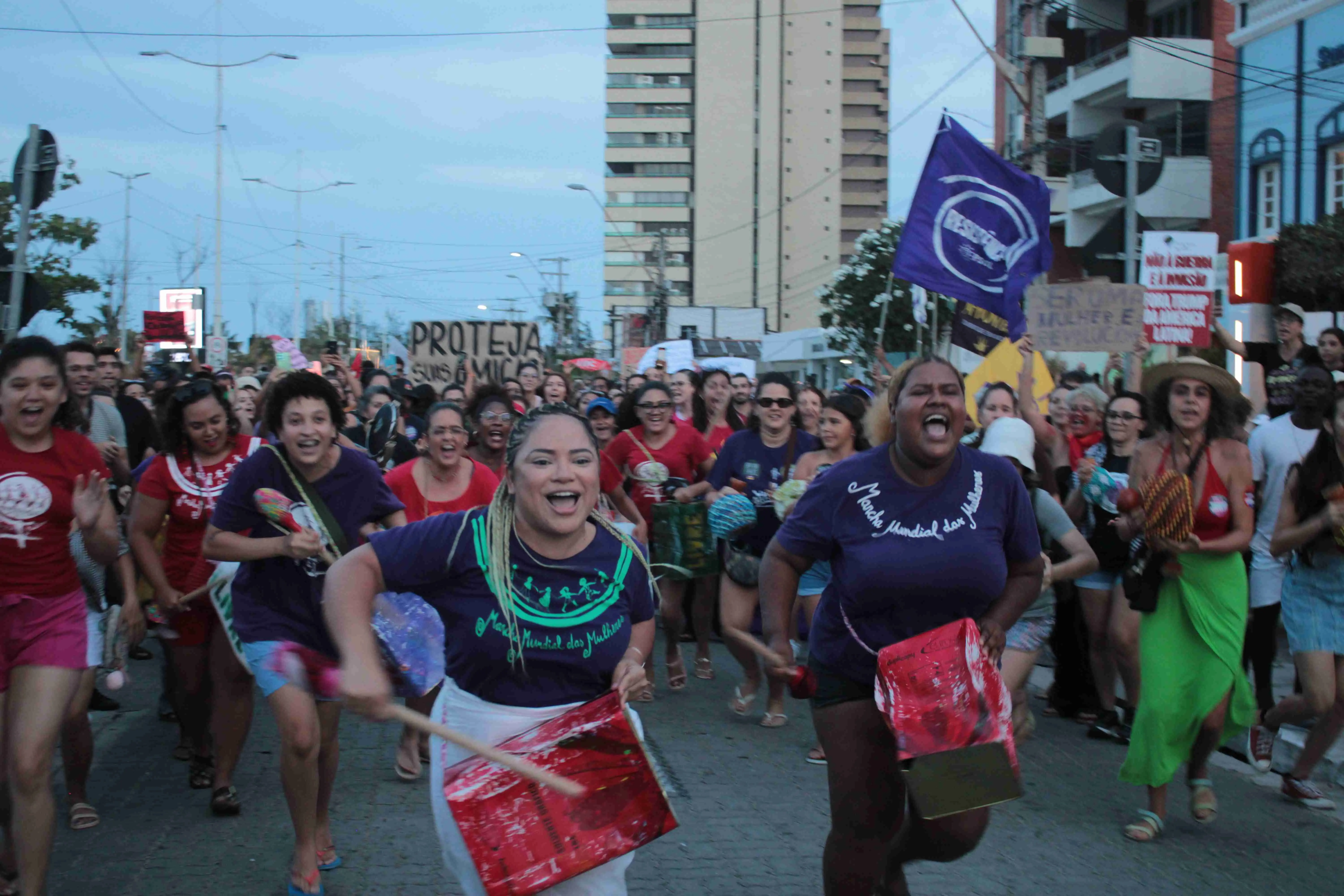 Um grupo de mulheres diversas, sorridentes e enérgicas, marchando em uma rua urbana, algumas tocando tambores e segurando cartazes de protesto.