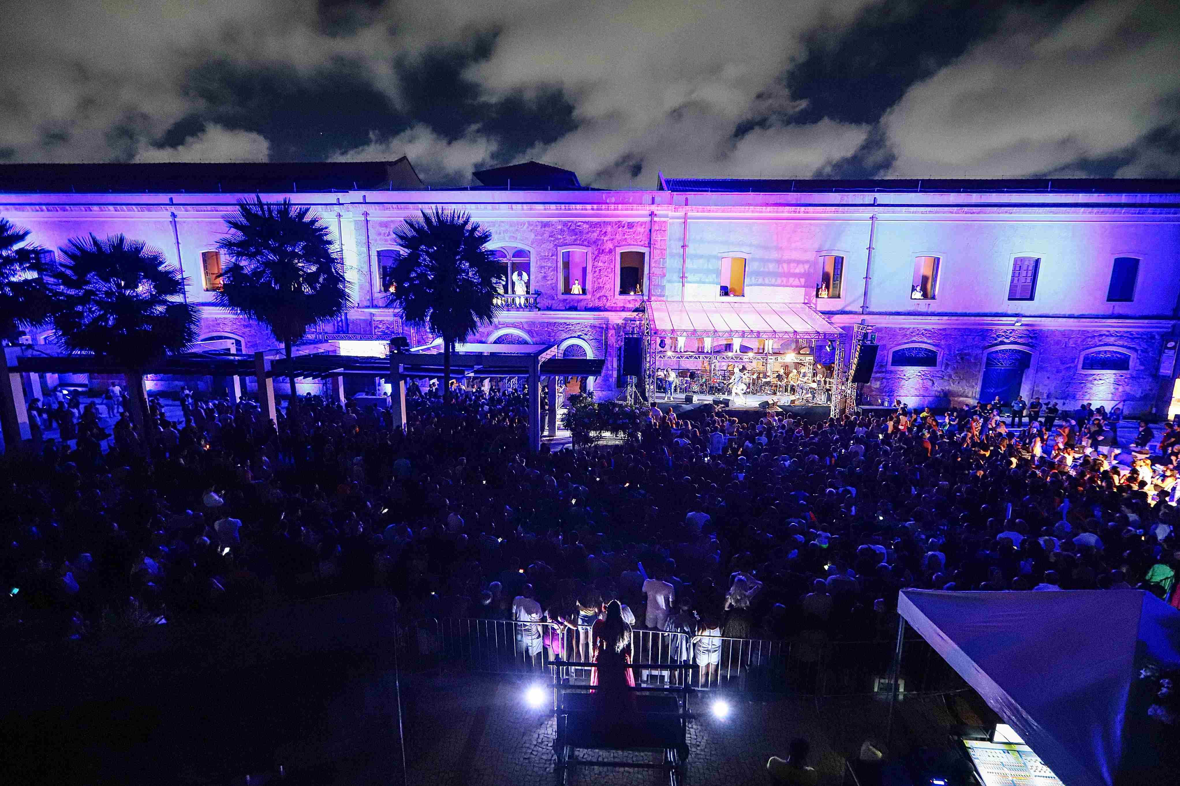 Uma fotografia noturna, tirada de cima, mostra um vibrante show ou evento ao ar livre em frente a um longo edifício histórico iluminado por uma forte luz roxa e azul. O palco, visível no centro da cena, está montado em frente à fachada do prédio, e a área logo abaixo está completamente lotada por uma grande e densa multidão. A iluminação dramática, que inclui as cores roxas e azuis refletidas no público e no edifício, junto com as silhuetas de palmeiras no lado esquerdo, cria uma atmosfera de festa noturna e celebração em larga escala.
