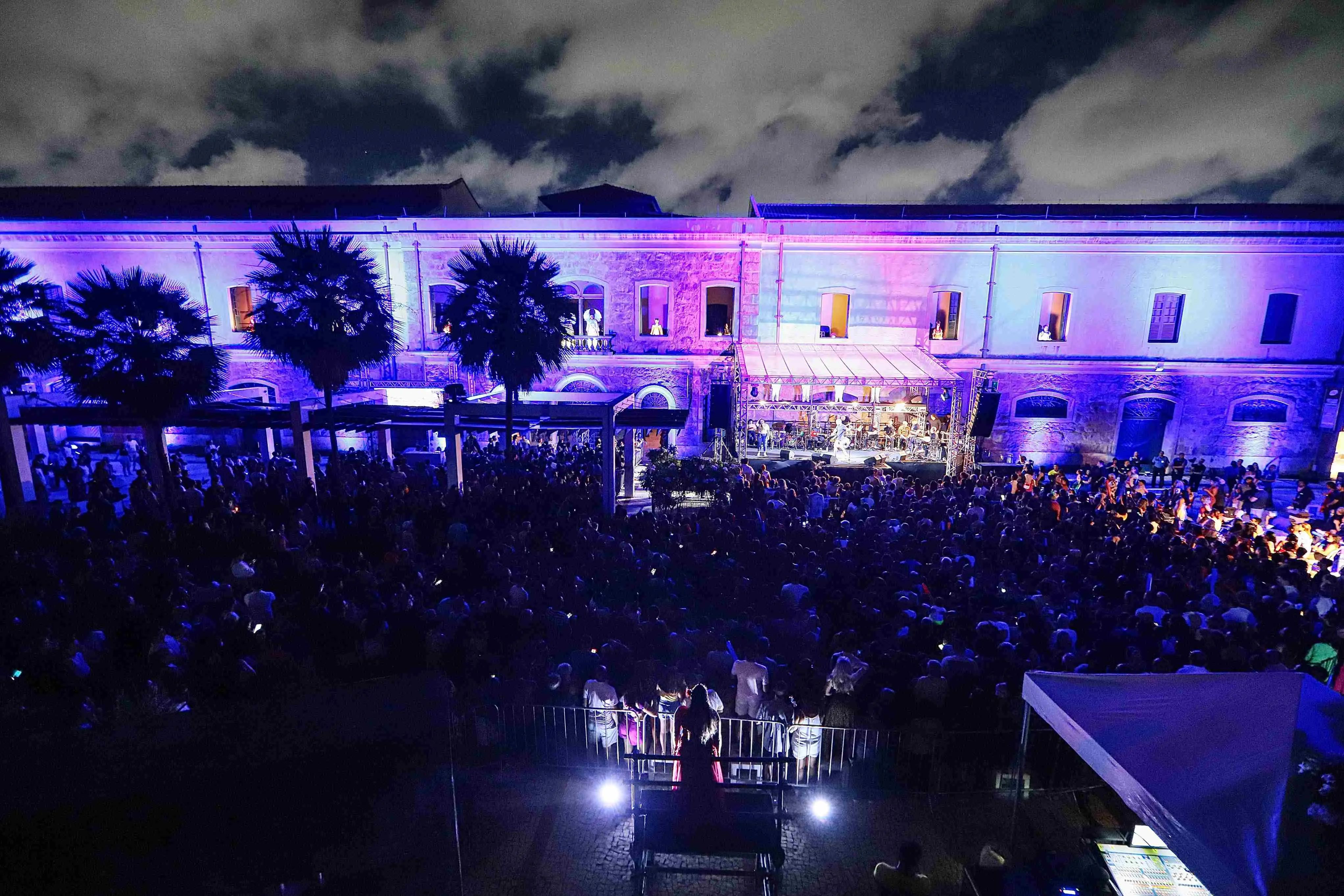 Uma fotografia noturna, tirada de cima, mostra um vibrante show ou evento ao ar livre em frente a um longo edifício histórico iluminado por uma forte luz roxa e azul. O palco, visível no centro da cena, está montado em frente à fachada do prédio, e a área logo abaixo está completamente lotada por uma grande e densa multidão. A iluminação dramática, que inclui as cores roxas e azuis refletidas no público e no edifício, junto com as silhuetas de palmeiras no lado esquerdo, cria uma atmosfera de festa noturna e celebração em larga escala.