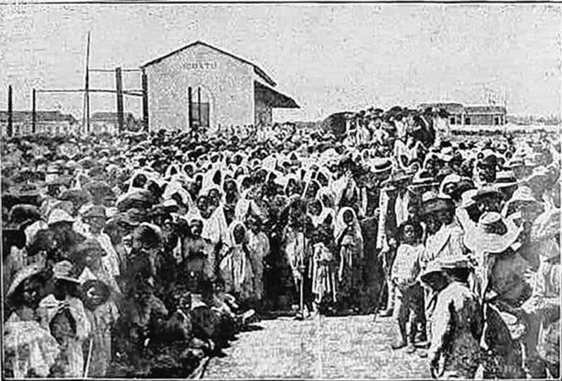 Uma foto em preto e branco mostra uma imensa multidão, possivelmente flagelados da seca, reunidas em um espaço aberto. A multidão é densa, com muitas pessoas vestindo roupas simples e chapéus. O alto número de pessoas sugere um acampamento, ponto de encontro ou local de auxílio, provavelmente relacionado a um período de grande seca e migração.