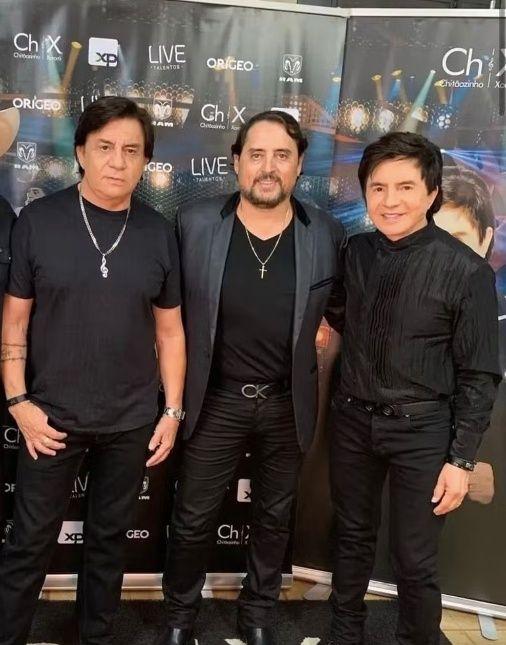 Três homens vestidos de preto, possivelmente um trio musical, posam em frente a um painel promocional com a marca 'Ch X Live'. Mauri era irmão de Chitãozinho e Xororó.