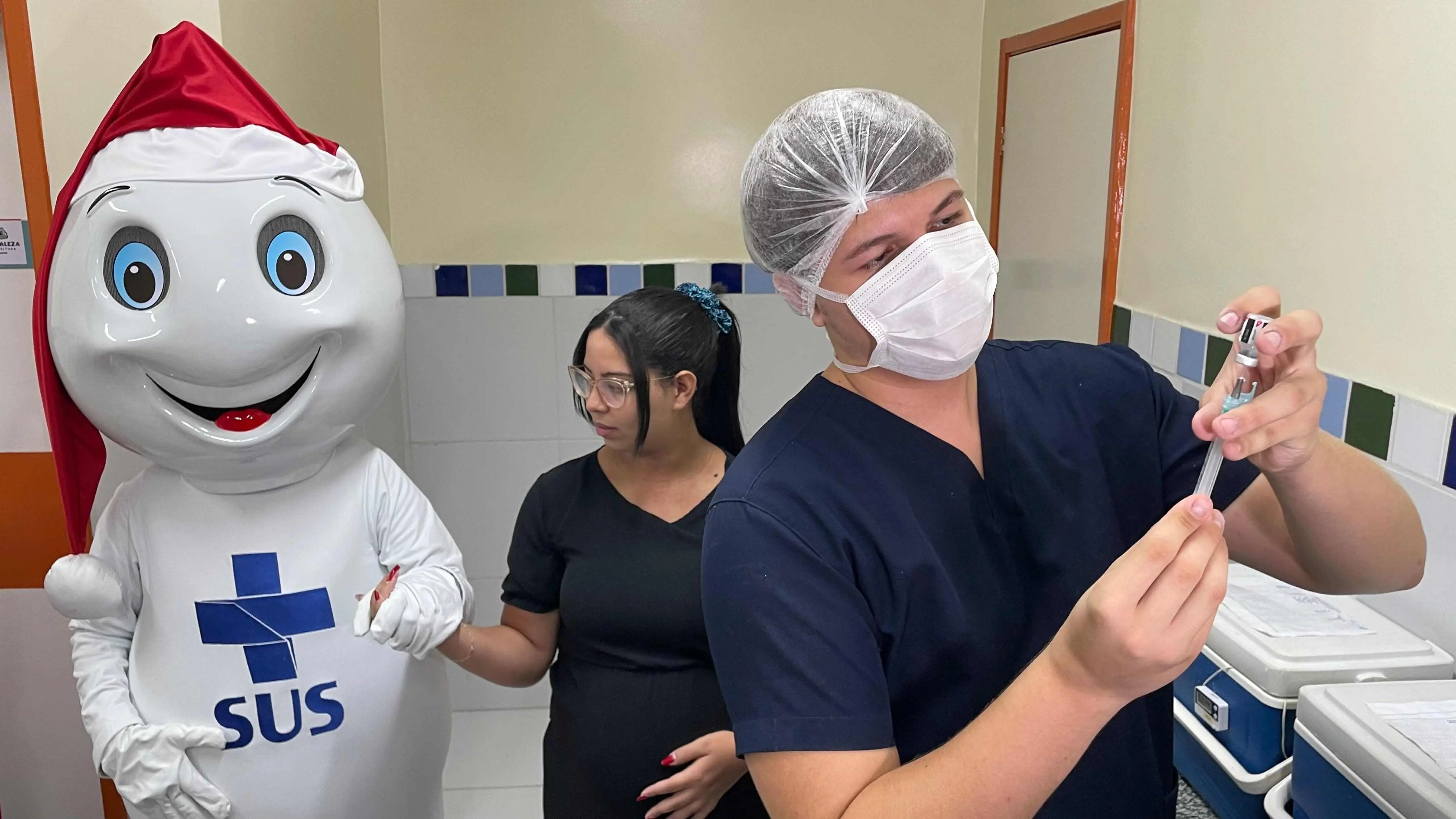 Profissional de saúde se preparando para aplicar vacina com a presença de um mascote do SUS vestido com gorro de Papai Noel, em ambiente hospitalar.