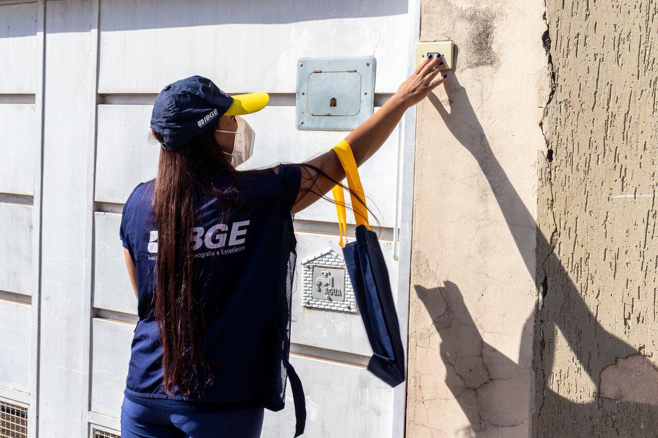 Mulher de cabelo longo liso e escuro, de costas e com máscara no rosto, tocando campainha de casa com portão branco. Ela usa blusa azul escura com nome IBGE e boné azul e amarelo.