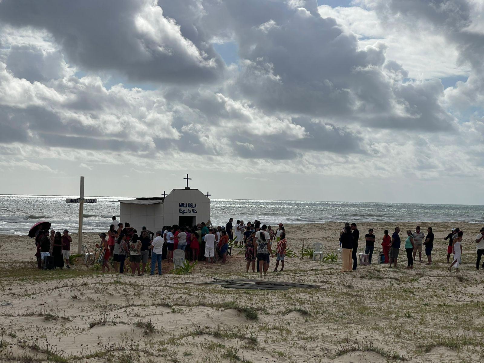 Vista panorâmica de uma pequena capela branca em uma praia arenosa, com o mar e o céu nublado ao fundo, e um grupo de pessoas reunidas ao redor.