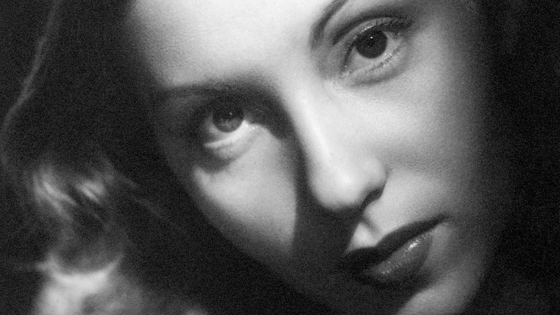 Na imagem, retrato em preto e branco e em close-up de Clarice Lispector. A imagem foca no rosto da escritora, em um ângulo que destaca seus olhos grandes e expressivos, um pouco voltados para cima e para a esquerda. A iluminação é dramática, com sombras fortes (chiaroscuro) que acentuam as maçãs do rosto e o contorno do nariz. Ela tem cabelos ondulados e escuros, e seus lábios estão levemente entreabertos, com um toque de batom. A foto transmite uma expressão de intensidade e mistério.