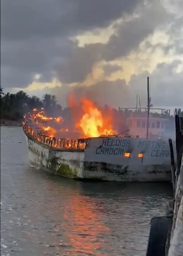 A imagem mostra um barco, de cor branca, sendo incendiado em alto mar. A embarcação tem escrito 'Heloísa Martins. Camocim, Ceará'.