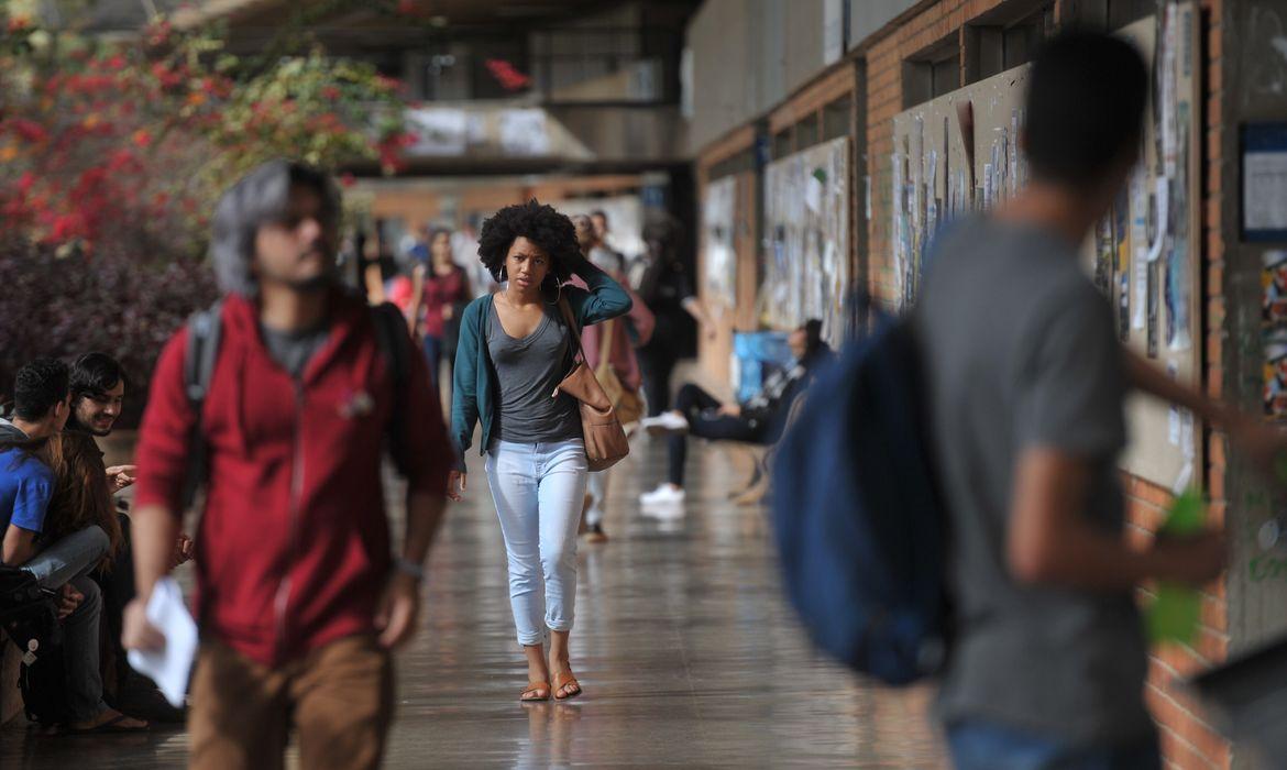Jovem mulher negra com cabelo crespo caminhando em um corredor movimentado de campus universitário, com outros estudantes e murais de aviso.