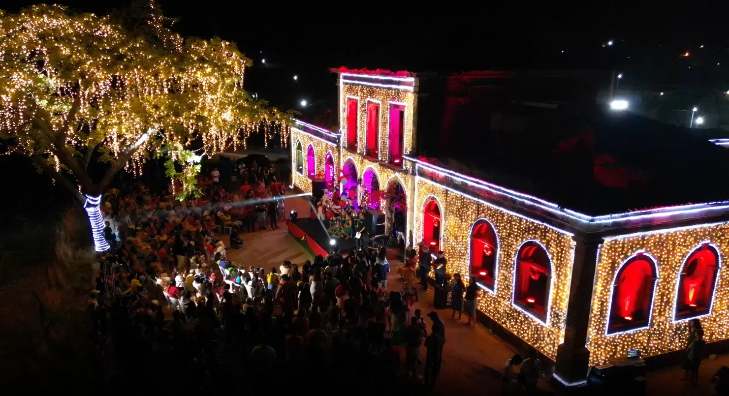 Foto aérea noturna de um casarão histórico ricamente iluminado. O edifício de fachada em arcos está contornado por luzes de Natal amarelas brilhantes, e as aberturas estão iluminadas em vermelho magenta. Uma grande árvore à esquerda também está coberta de luzes tipo cascata. Uma multidão está reunida em frente à construção, celebrando um evento noturno.