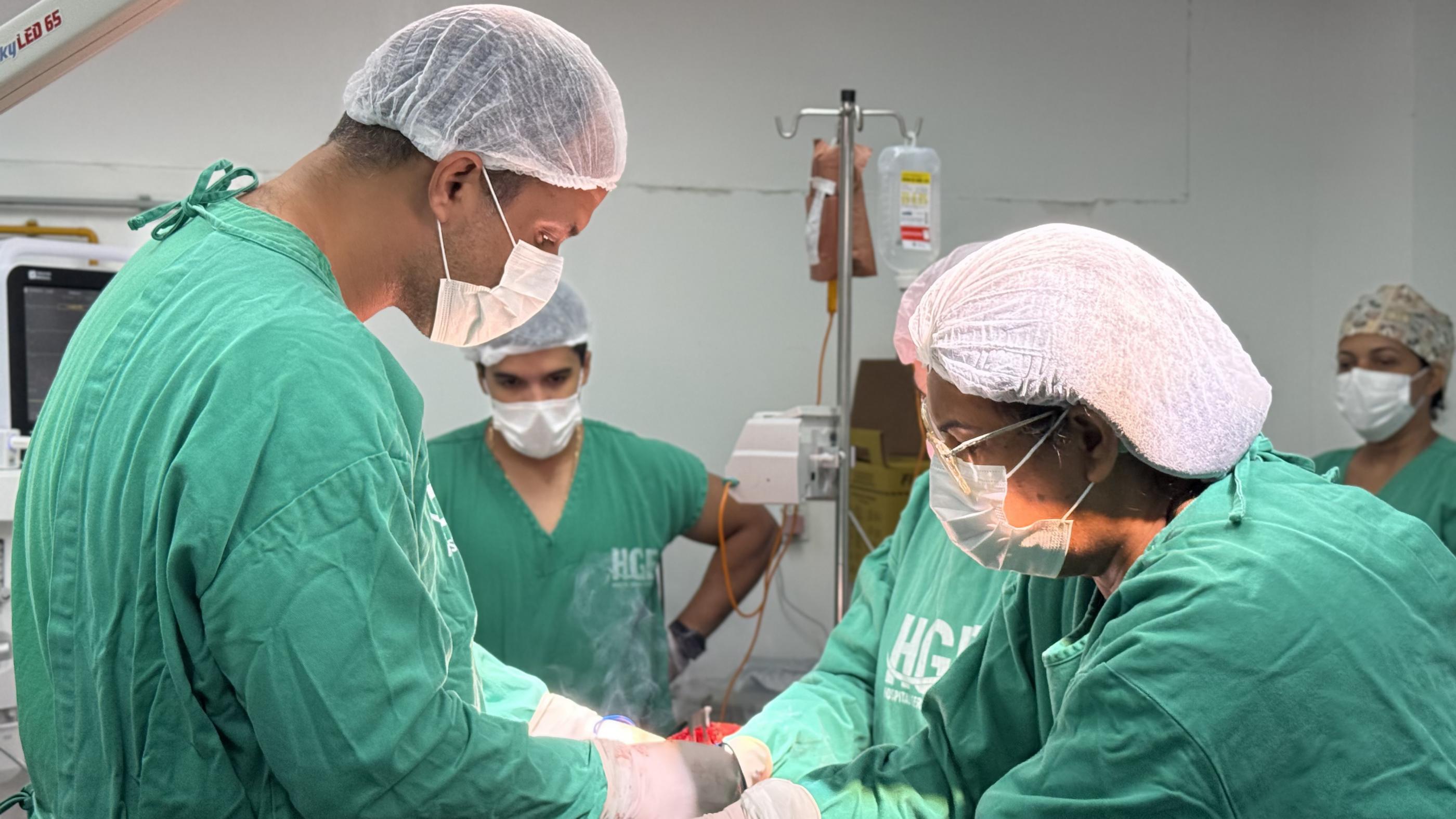 Equipe médica realiza procedimento cirúrgico em uma sala de operação. Profissionais vestem aventais verdes, toucas descartáveis e luvas estéreis. No centro, há uma mesa cirúrgica com instrumentos e área coberta por campos cirúrgicos. Ao fundo, é possível ver um suporte com bolsa de soro e equipamentos hospitalares.