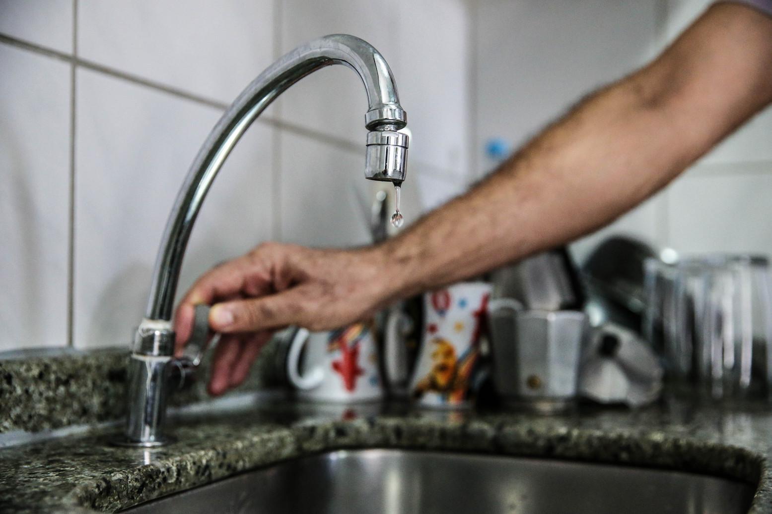 Mão de uma pessoa fechando uma torneira de cozinha cromada, da qual cai uma gota d'água, sobre uma pia de inox e bancada de granito.