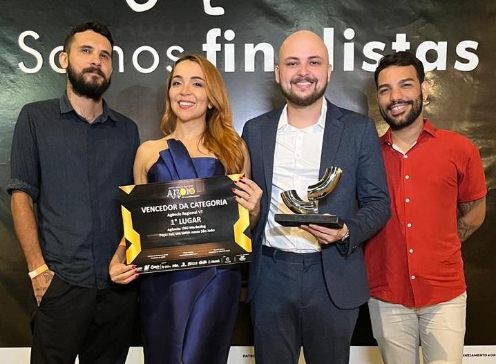 A imagem mostra quatro pessoas posando diante de um painel de premiação. No centro, duas delas seguram um troféu e uma placa que indica a conquista de um prêmio na categoria em que participaram. Todos estão bem vestidos e sorriem para a câmera