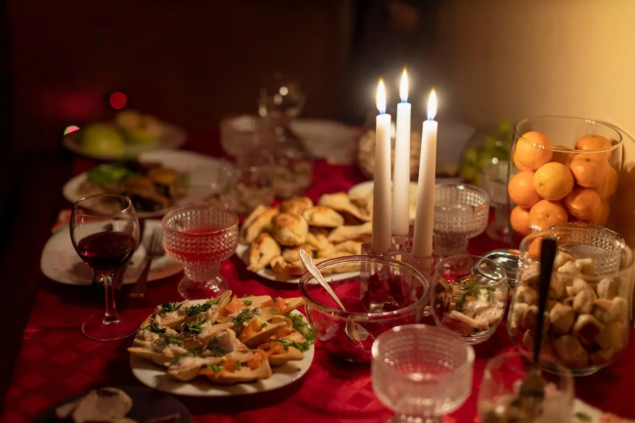 Mesa de toalha vermelha repleta de variados pratos de comida e taças de vinho. Ao centro, três velas.