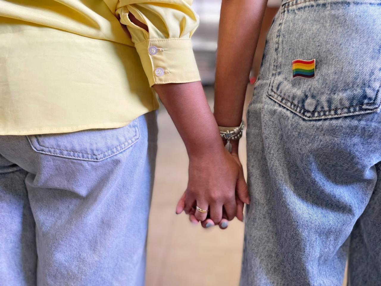 Duas mulheres segurando as mãos, uma com camisa amarela e a outra com um pin da bandeira LGBTQ+ no bolso da calça jeans clara.