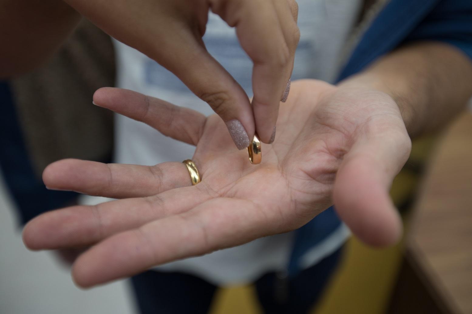 Mão de mulher com unhas pintadas colocando aliança dourada na palma de outra mão masculina, com outra aliança já no dedo anelar. A foto representa uma separação de casal.