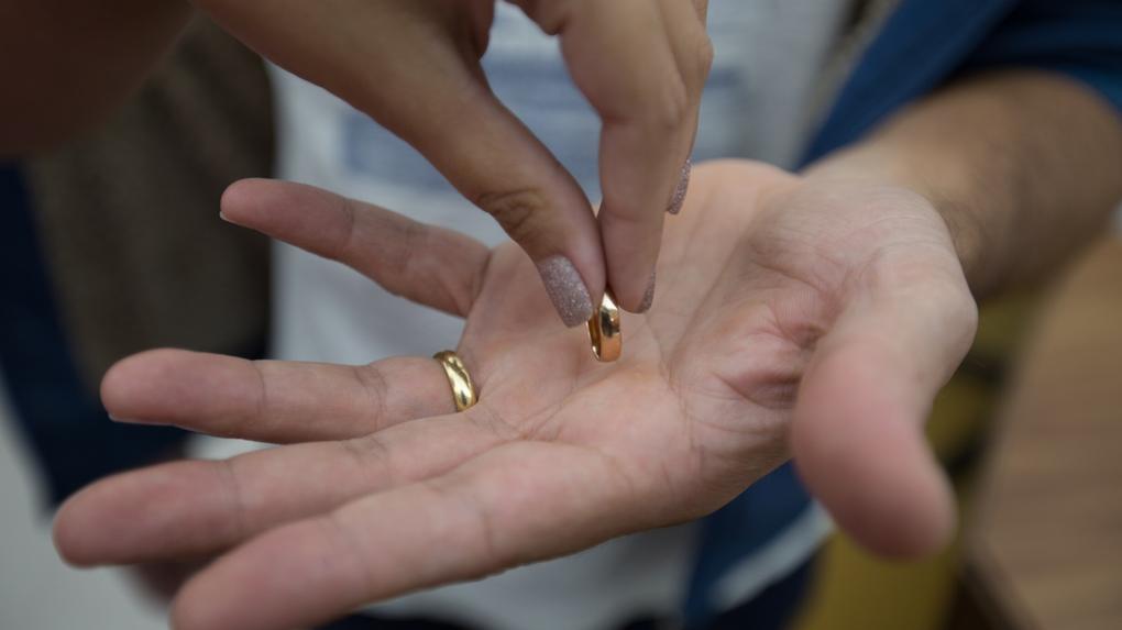 Mão de mulher com unhas pintadas colocando aliança dourada na palma de outra mão masculina, com outra aliança já no dedo anelar. A foto representa uma separação de casal.