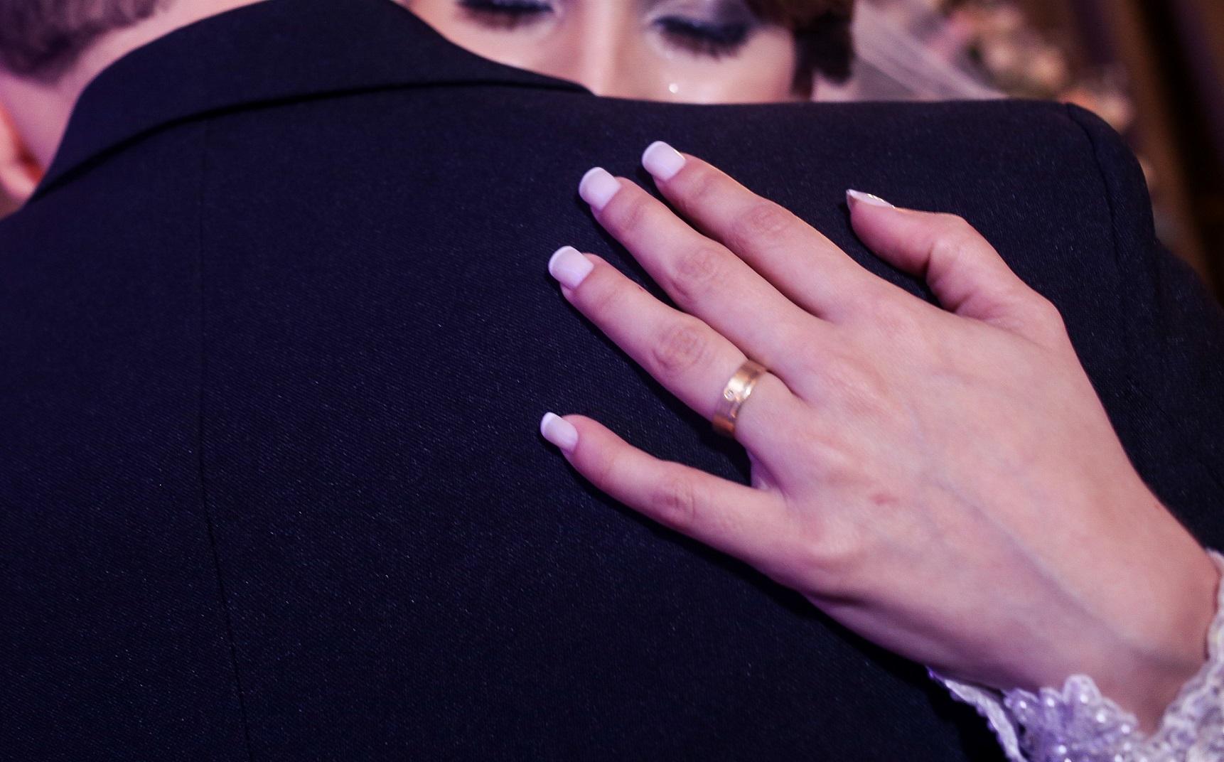 Close-up da mão de uma noiva com anel de ouro no dedo anelar, abraçando o marido, em uma festa de casamento. O rosto dela aparece desfocado ao fundo.