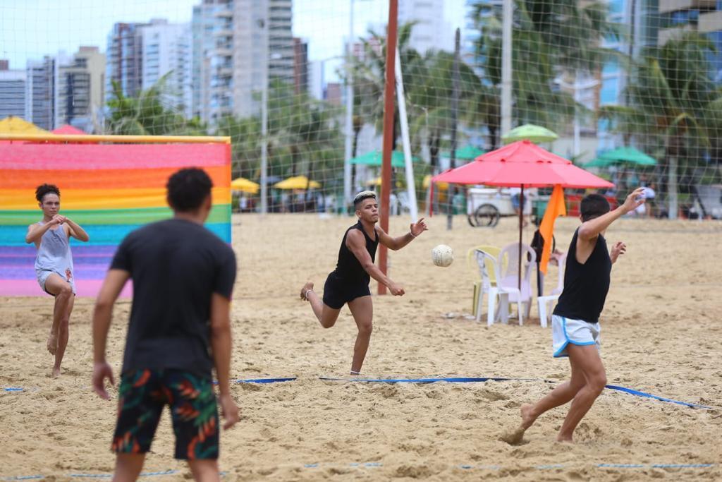 Na imagem, quatro pessoas jogam futevôlei em uma quadra de areia na praia. Um jogador no centro, vestindo regata preta e shorts preto, está saltando para golpear a bola com o pé. Outros dois jogadores estão à direita e à esquerda. Ao fundo, há prédios, palmeiras e guarda-sóis coloridos.