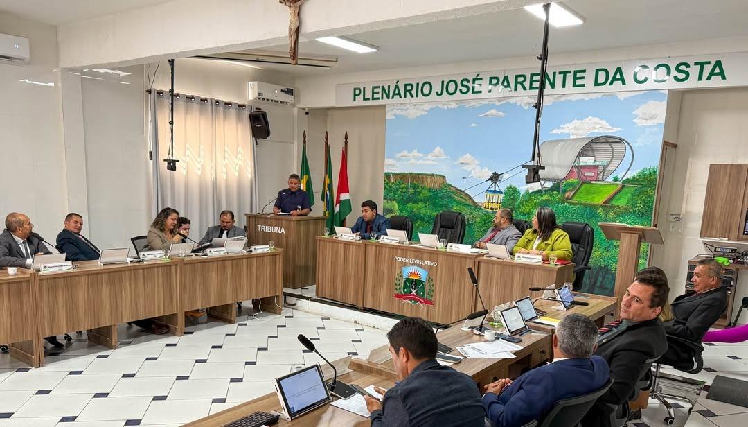 Sessão da Câmara Municipal em plenário com vereadores reunidos em torno de mesas em formato de U. Um homem fala na tribuna. Ao fundo, mural colorido com ilustração do teleférico de Ubajara e paredes com o nome ‘Plenário José Parente da Costa’. Vários vereadores utilizam notebooks durante a sessão.