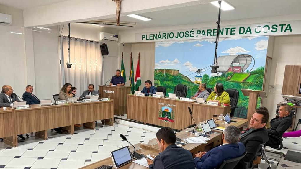Sessão da Câmara Municipal em plenário com vereadores reunidos em torno de mesas em formato de U. Um homem fala na tribuna. Ao fundo, mural colorido com ilustração do teleférico de Ubajara e paredes com o nome ‘Plenário José Parente da Costa’. Vários vereadores utilizam notebooks durante a sessão.